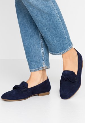 Navy blue suede loafers with a bow detail, flat tan sole, and stitched accents, paired with cropped light blue jeans.