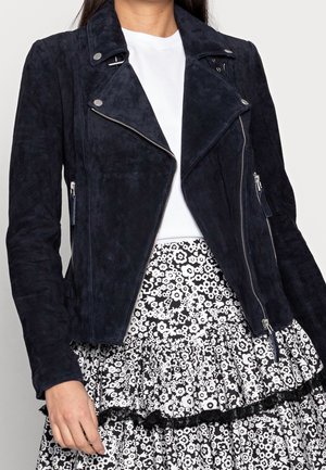 Femme portant une veste biker en daim noir sur un top blanc et une jupe à motif floral noir et blanc avec des volants.