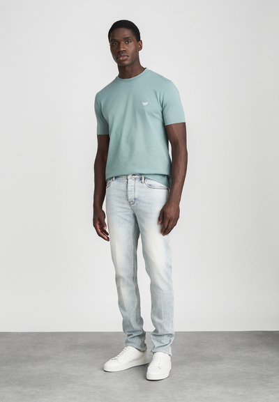 Mann som står mot en ensfarget bakgrunn iført en lyseblå kortermet skjorte, lyse blå jeans og hvite sneakers.