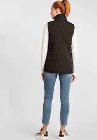 Svart vadderad ärmlös väst med hög krage, bakstycken och en sl�ät textur, kombinerad med ljusblå skinny jeans och sneakers.