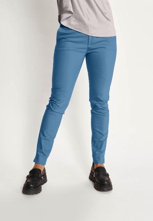 Person trägt enganliegende blaue Hosen und schwarze klobige Loafers und steht vor einem schlichten hellen Hintergrund.