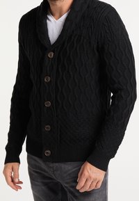 Cardigan nero lavorato a maglia con motivi testurizzati, colletto a scialle e cinque bottoni marroni, abbinato a jeans grigio scuro.