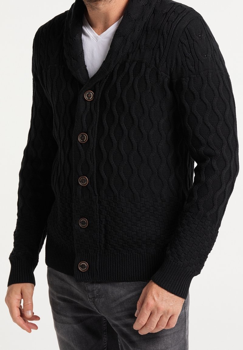 Cardigan nero lavorato a maglia con motivi testurizzati, colletto a scialle e cinque bottoni marroni, abbinato a jeans grigio scuro.