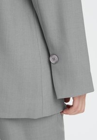 Lysegrå blazer med tekstureret stof, der har en bred manchet og en rund lilla knapdetalje på ærmet.