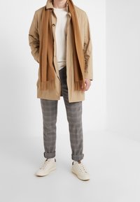 Beige lättviktig kappa över en krämfärgad skjorta, parat med grå rutiga byxor och vita sneakers, kompletterat med en brun fransad scarf.