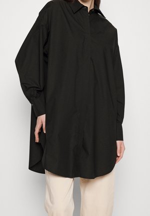 Frau trägt ein übergroßes schwarzes Hemd mit langen Ärmeln und beigen Hosen vor einem einfarbigen Hintergrund.