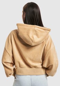 Hoodie en velours côtelé beige coupé, avec une surface texturée, comportant une capuche et des manches larges. Détail de taille élastique visible.