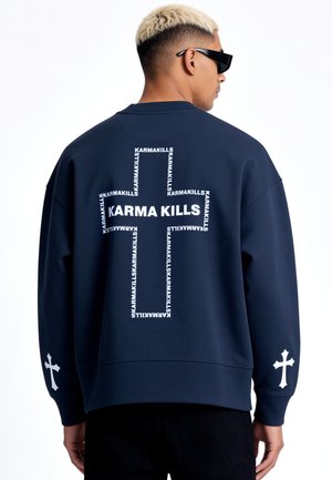 Homme aux cheveux blonds courts et lunettes de soleil noires portant un sweat bleu marine avec des motifs de croix blanches et le texte "KARMA KILLS" au dos et sur les manches.