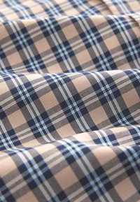 Tissu à carreaux présentant un motif écossais beige et bleu marine avec des accents bleu clair. La texture semble lisse et tissée.