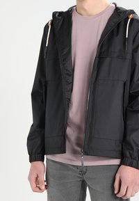 Zwarte lichtgewicht jas met een ritssluiting aan de voorkant, twee voorzakken, verstelbare capuchon met trekkoorden en elastische manchetten. Gedragen over een lichtroze shirt.