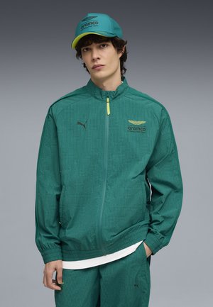 Junger Mann mit grünem Aramco Formula One Team-Jacke und passender Kappe, der mit einer Hand in der Tasche vor grauem Hintergrund steht.