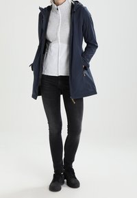 Marineblaue, wasserdichte Jacke mit Kapuze, vorne mit Reißverschluss und Taschen, darüber ein hellgraues Fleece-Oberteil und schwarze Jeans.