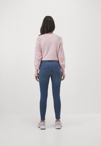 Világos rózsaszín pulóver, kötött mintás ujjakkal, sötétkék skinny farmerral párosítva. A lábbeli fehér, rózsaszín kiegészítőkkel. Egyszerű dizájn, logók nélkül.