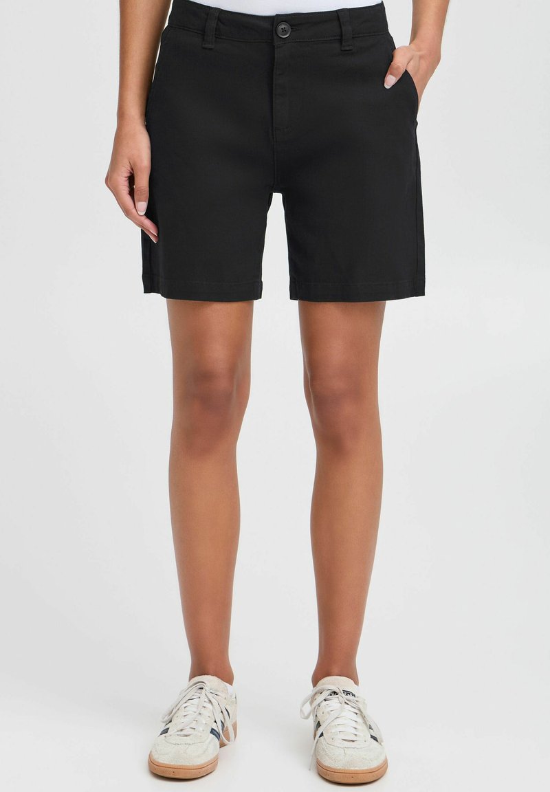 Sorte shorts i bomullsmiks med en tettsittende midje, sidelommer og rett passform, kombinert med hvite sneakers på en lys bakgrunn.