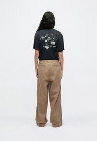 Oseba z dolgimi temnimi lasmi, oblečena v črno majico z risbami pijač in bež hlače širokih hlačnic, stoji obrnjena proti svetlemu ozadju brez vzorcev.