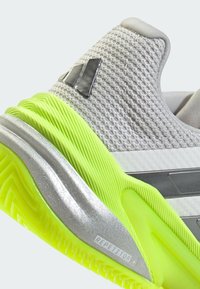 Zapatilla deportiva gris con parte superior de malla texturizada, suela de color amarillo neón, detalles en plata y un logo en el lateral. Patrón de suela estriado visible.