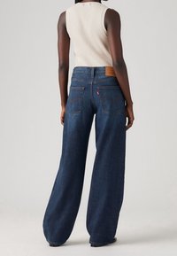 Kvinna i vida, mörkblå denimjeans och en ärmlös kremfärgad ribbad topp, stående mot en enfärgad bakgrund, med ryggen vänd mot kameran.