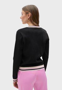 Crna dukserica sa okruglim izrezom, rebrastim manžetama i prugastim rubom s roze, bijelim i zlatnim akcentima. Model nosi roze hlače.