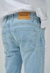 Vaqueros de denim azul claro con una etiqueta de cuero en la parte trasera que dice "Salsa Jeans", con detalles de costura y diseño estándar de cinco bolsillos.