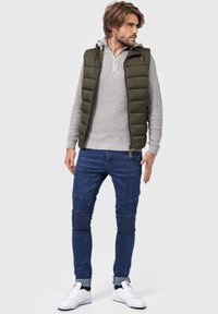 Polstret olivengrøn vest, grå ribbet sweater, blå slim-fit jeans med ribbede detaljer og hvide sneakers. Lækkert lag-på-lag outfit.