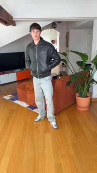 Sort skinn bomberjakke med glidelås foran, lyseblå jeans og sølvfargede joggesko. Bakgrunnen har en plante og en hjørnesofa.