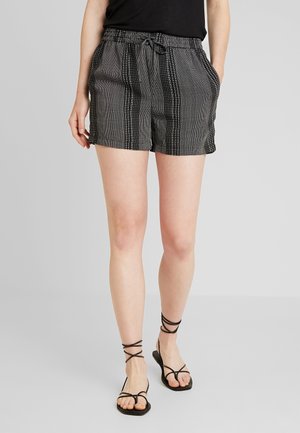 Frau trägt schwarz-weiß gestreifte Shorts mit Kordelzug, schwarzes Top und schwarze Riemchensandalen und steht vor einem schlichten Hintergrund.