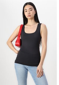 Schwarzes, enges Tanktop mit Rundhalsausschnitt, getragen mit hellblauen Jeans. Eine rote Ledertasche hängt über der rechten Schulter. Glatte Stofftextur.