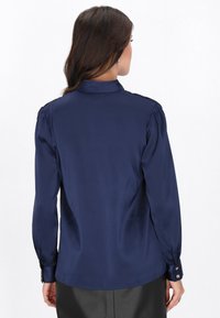 Mørkeblå bluse med lange ærmer, høj krave og puffede skuldre. Glat stof med knaplukning ved ærmerne og en skræddersyet pasform.