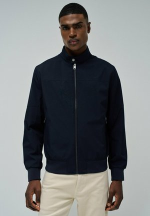 Chaqueta de zip azul marino con cuello alto, puños acanalados y dos bolsillos frontales, que presenta una textura suave y un diseño minimalista.