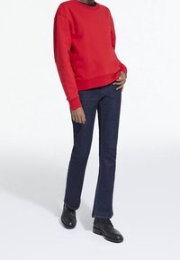 Sudadera roja con cuello redondo y puños de canalé, combinada con jeans acampanados oscuros y botas negras de tobillo, con una textura suave.