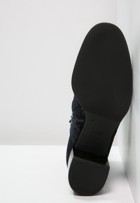 Suela de goma negra de un zapato, con una superficie texturizada. Parte superior hecha de tela oscura con un diseño intrincado. Logo de la marca visible.