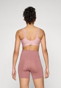 Sostén deportivo de color rosa claro y pantalones cortos de cintura alta con una textura suave. Marca visible en las correas. Diseño simple y atlético.