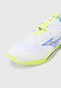 Fehér sportcipő neon sárga kiemelésekkel, texturált hálós felsőrésszel és kék díszítéssel az oldalon. Kerek orr és többszínű talp.