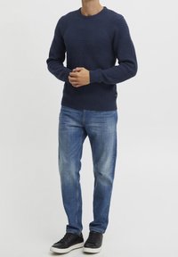 Marineblauer Strickpullover mit strukturiertem Muster im oberen Bereich, gerippten Bündchen und Saum, kombiniert mit hellblauen Jeans und schwarzen Sneakern.