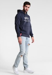 Marineblauwe hoodie met een logo aan de voorkant, een buidelzak en een capuchon met trekkoord, gecombineerd met lichtblauwe ripped jeans en witte sneakers.