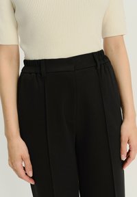 Schwarze maßgeschneiderte Hose mit glatter Textur, ausgestattet mit einem elastischen Bund und vorderen Falten, kombiniert mit einem eng geschnittenen, gerippten cremfarbenen Oberteil.