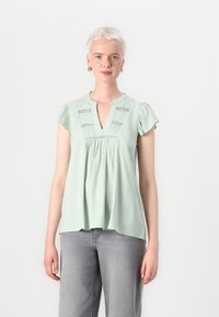 Blusa verde menta con maniche corte a volant, scollo a V e dettagli plissettati. Realizzata in tessuto morbido con una vestibilità comoda.