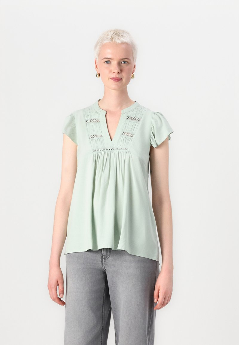 Blusa verde menta con maniche corte a volant, scollo a V e dettagli plissettati. Realizzata in tessuto morbido con una vestibilità comoda.