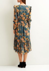 Abito midi floreale in teal con maniche lunghe, polsini arricciati e orlo a più strati. Presenta una vita aderente e un vivace motivo floreale in colori caldi.