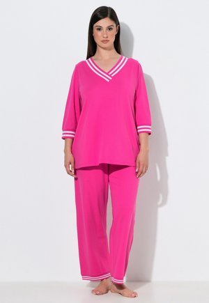 Vrouw staat op blote voeten en draagt een felroze set met lange mouwen top en broek met witte gestreepte biezen op mouwen, halslijn en broekspijpen.