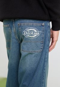 Blå denimbukser med en afslappet pasform, der har et hvidt broderet "Dickies" logo på baglommen og solide syninger.