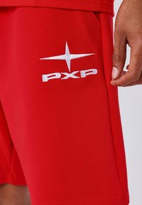 Shorts rouges avec un logo étoile brodé blanc et le texte "PXP" sur la cuisse, à côté de la main d'une personne reposant à ses côtés.