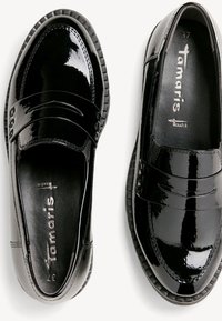 Mocassins en cuir verni noir avec un petit talon, un bout arrondi et une bride décorative sur le dessus, affichant le nom de la marque "Tamaris" à l'intérieur.