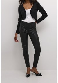 Schwarze, kurze Strickjacke über einem weißen Oberteil, kombiniert mit schwarzen Kunstlederhosen. Das Modell trägt schwarze High-Heel-Mules mit dekorativen Akzenten.