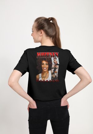 Kvinde med brunt hår i en hestehale iført en sort Whitney Houston grafisk t-shirt, der viser flere portrætter og hendes navn i røde bogstaver.