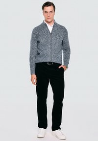 Szary dzianinowy sweter zapinany na zamek z teksturowanym wzorem, w połączeniu z czarnymi spodniami z korduroju i białymi sneakersami. Model stoi na neutralnym tle.