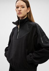 Femeie cu păr drept castaniu, purtând un pulover negru Calvin Klein cu fermoar pe jumătate și cu mâinile în buzunarele din față, pe un fundal simplu.