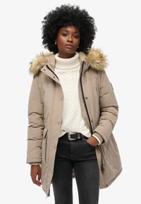 Parka beige con cappuccio rifinito in pelliccia, con chiusura a zip e bottoni. Indossata sopra un maglione bianco a maglia e jeans scuri.