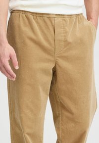 Cordhose in einem hellbraunen Farbton, ausgestattet mit einem strukturierten Streifenmuster, einem elastischen Bund und seitlichen Taschen für zusätzliche Bequemlichkeit.