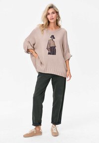 Beżowy, luźny sweter z dzianiny o teksturze ściągacza i graficznym nadruku, w połączeniu z ciemnozielonymi spodniami o luźnym kroju oraz beżowymi trampkami.